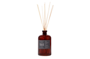 A reed diffuser with amber glass bottle and liquid, multiple light brown reeds inserted, label on front with product information.