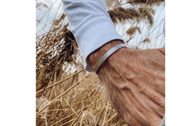 A close-up of a person's hand and wrist wearing a thin silver-toned metal cuff bracelet, set against a background of dry golden reeds.