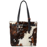 A structured cowhide tote bag with black leather shoulder straps and trim, featuring a brown and white natural cow print pattern and copper-toned hardware, shown against a plain white background.