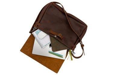 A brown leather shoulder bag shown open with a notebook, pen, and small pouch peeking out, photographed on a clean white background.